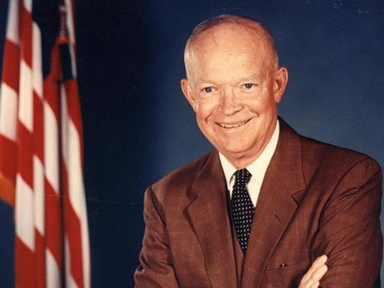 Dwight Eisenhower, America's 34th president. Smiling in the picture with folded arms, with the American flag in the background.