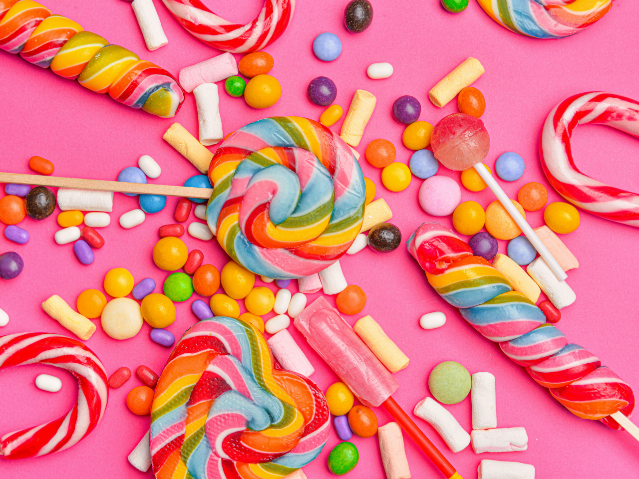 Colourful sweets such as candy canes, lollipops, jellybeans, and marshmallows spread out over a pink, flat surface
