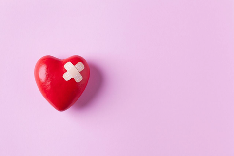 red heart with white band aid in a x shape on a pink background