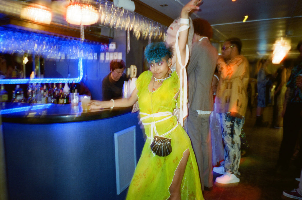 model posing at bar with left arm up, person making drink behind.