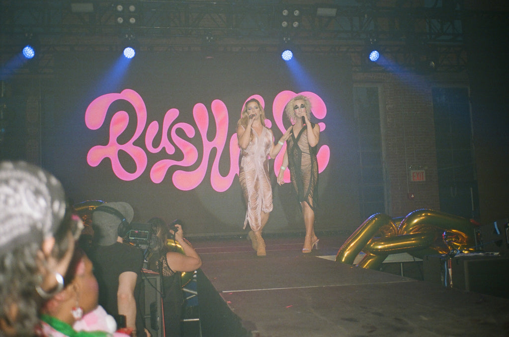 hosts in front of bushwig sign on stage.