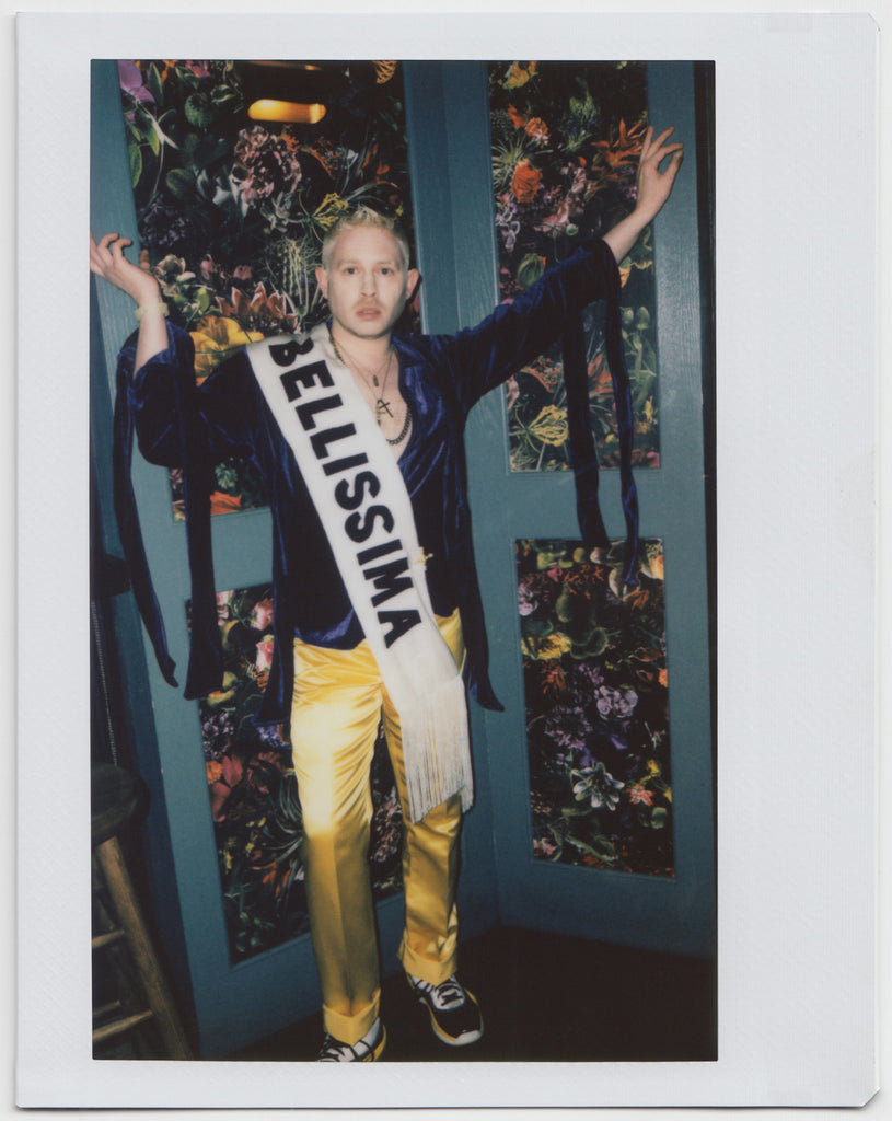 person posing wearing sash with printed 'bellissima' text, arms raised, looking directly at the camera - polaroid style.