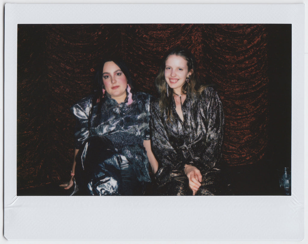 duo wearing silver outfits posing to the camera, velvet background - polaroid style. 