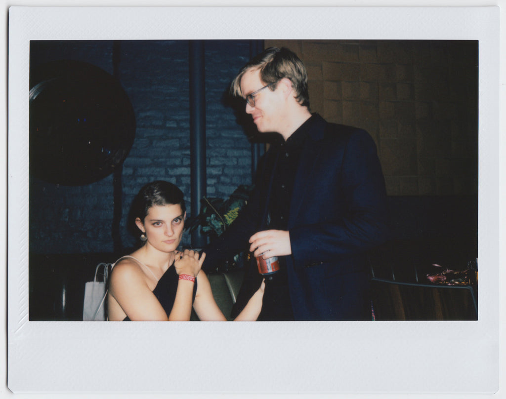 two people posing: one with hand on other's shoulder and holding a drink, the other looking directly at the camera - polaroid style. 