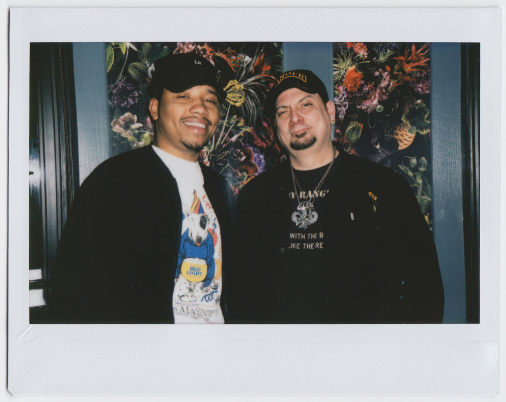 two people wearing hats, smiling side by side with floral wallpaper backdrop - polaroid style.