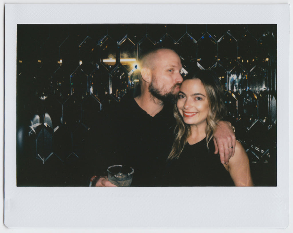 duo posing, one person holding a drink and kissing the other's head, mirrored wall in the background - polaroid style. 