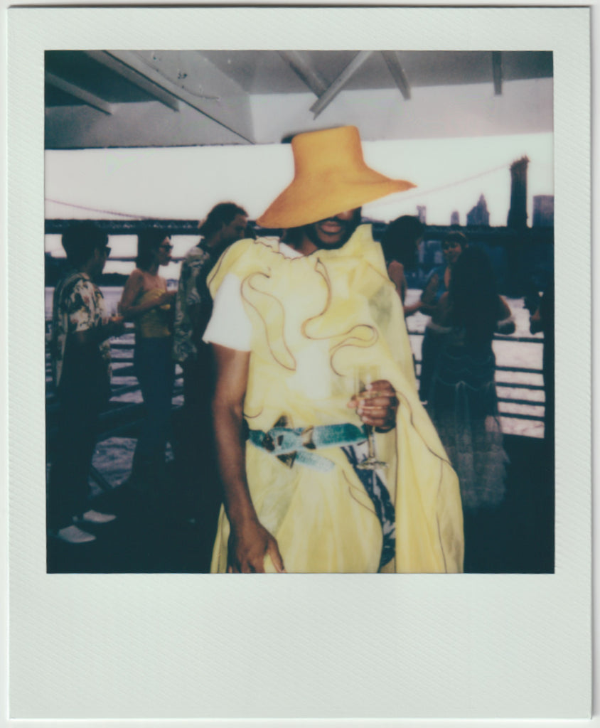 model in dress with hat covering face, people standing behind.
