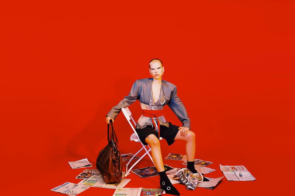 model seated on chair surrounded by news paper and holding a bag.