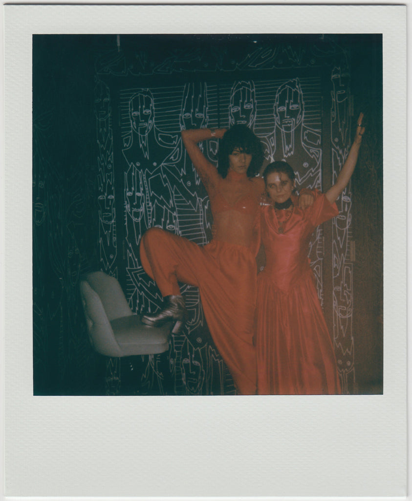 duo posing in all-red outfits, hands raised, one of the people with their leg on a chair - polaroid style.