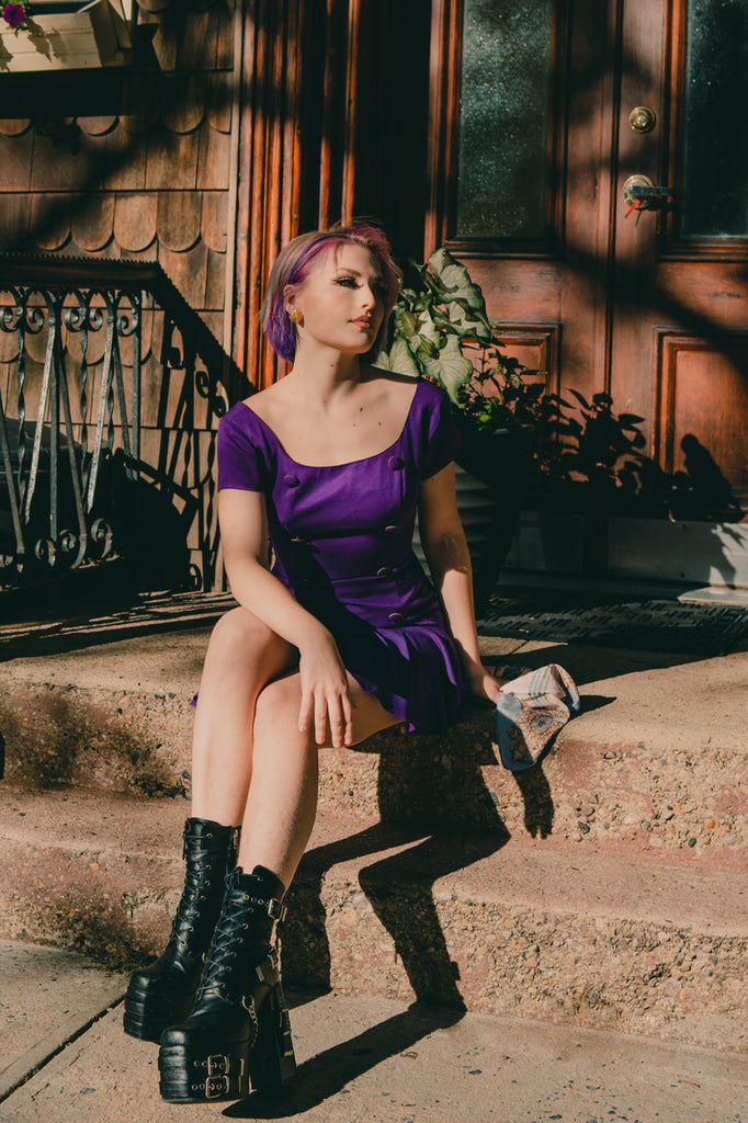 sofia coll in a purple dress and black chunky boots seated on stoop 