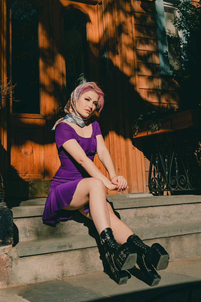 sofia coll in a purple dress and black chunky boots seated on stoop 