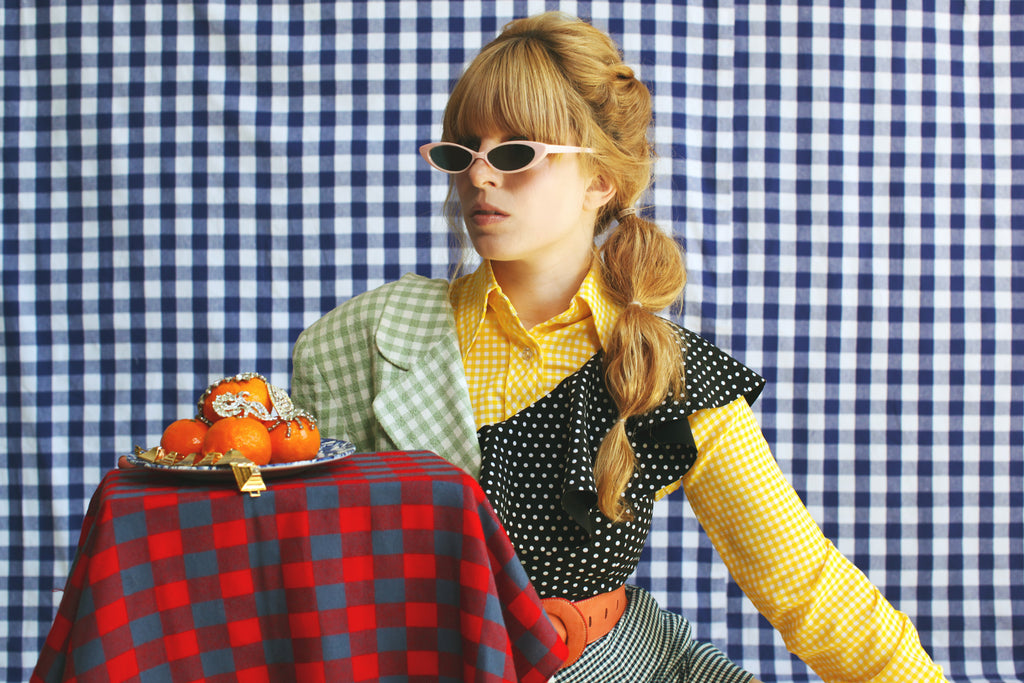 model in slim sunglasses mixed check prints and polka dots in front of a checked backdrop with a plate and oranges still life.