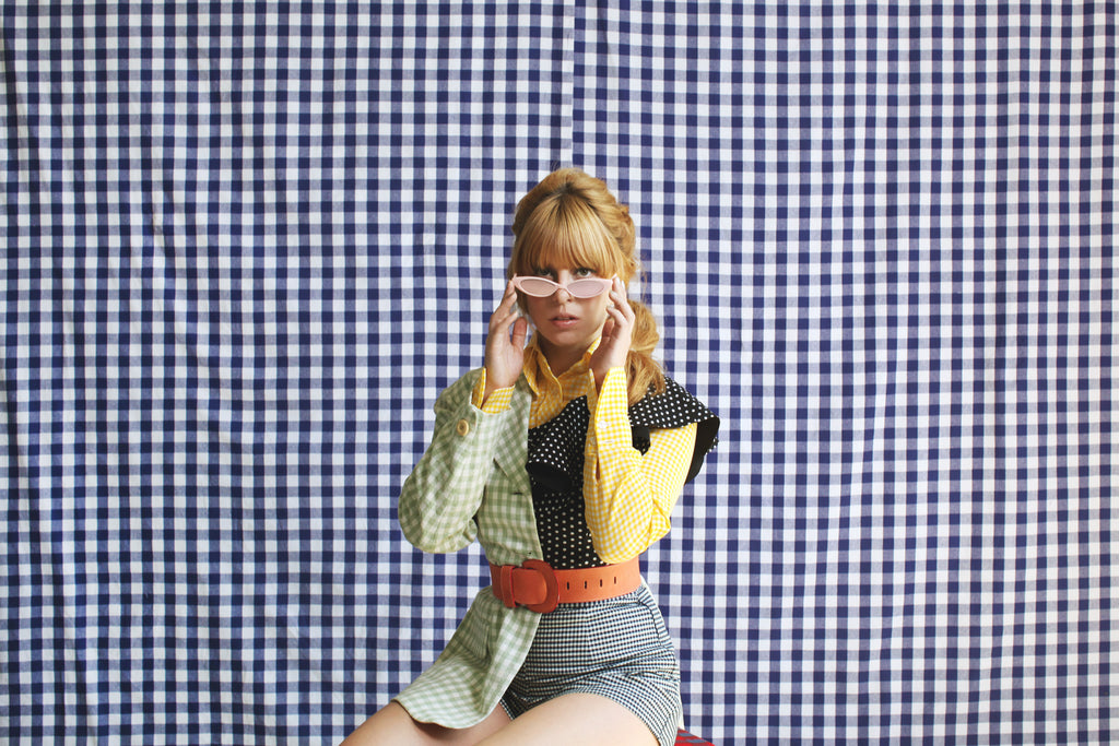 model seated in shorts, slim sunglasses mixed check prints and polka dots in front of a checked backdrop .