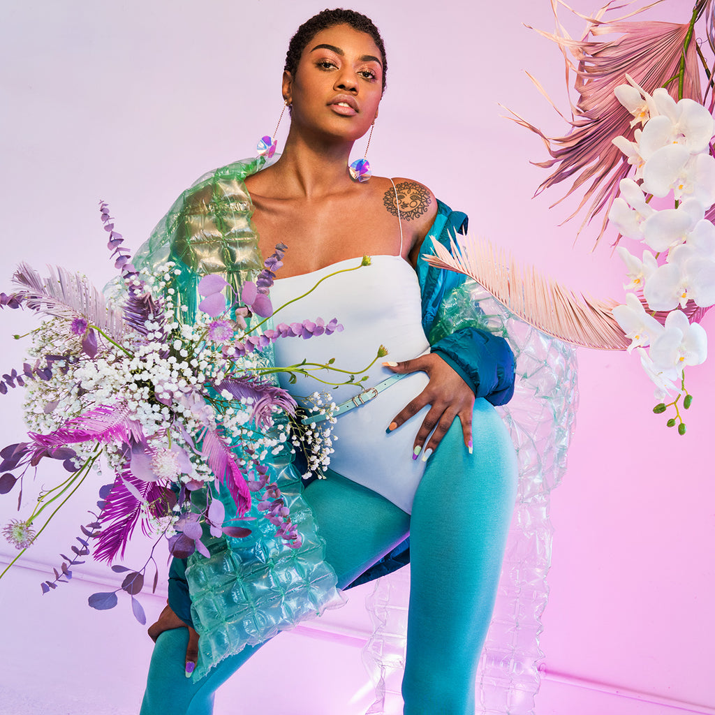 single model posing in body suit, tights, a puffy jacket and bubble wrap surrounded by floral displays. 