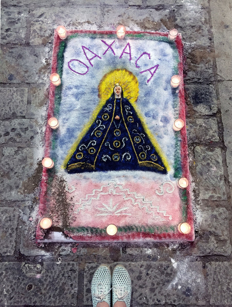 top view of sand art with feet, a saint and the word 'oaxaca', framed by candles on an old street.
