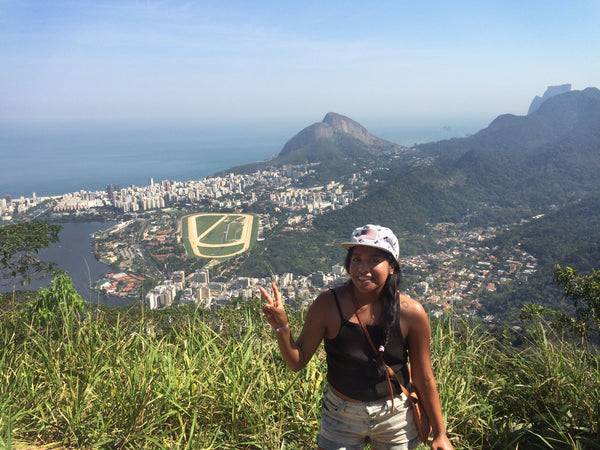 person smiling at the camera, making a peace and love gesture, at the top of corcovado mountain with a view of rio de janeiro in the background.