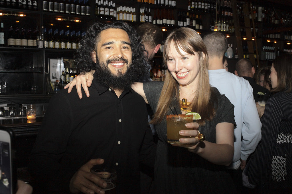 duo posing with drinks, bar in the background.