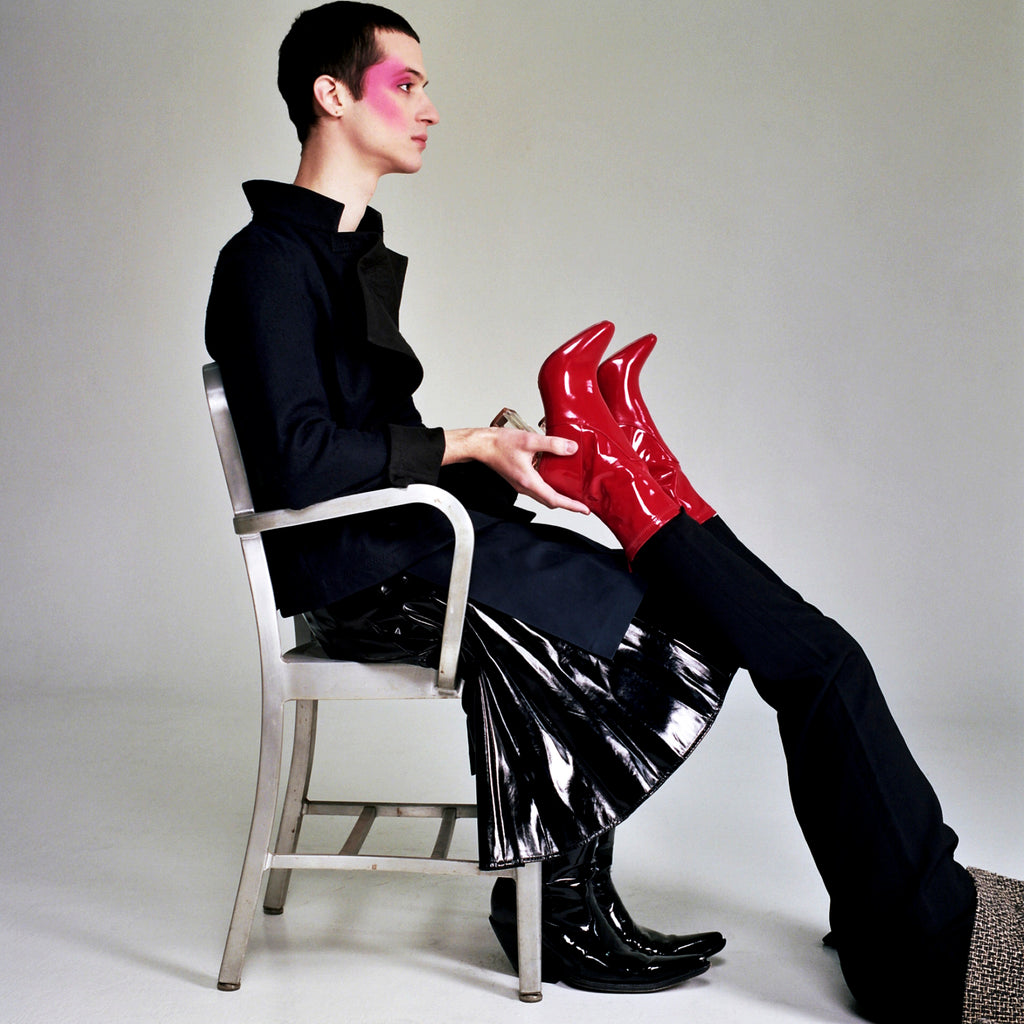 one model seated in chair holding up red boots of other model laying at their feet.