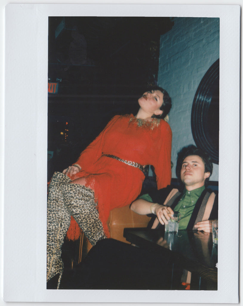 two people posing: one sitting at a table with a drink, the other at an elevated level, both looking directly at the camera - polaroid style.