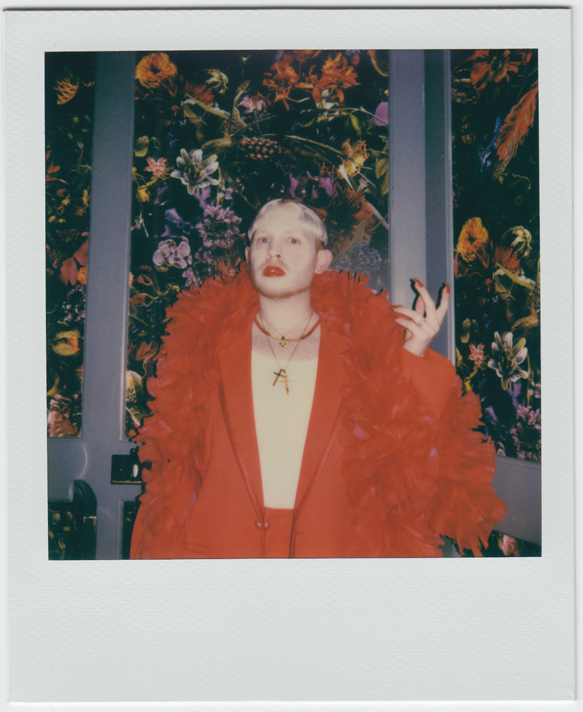 dressed all in red, employee poses with left hand raised, floral pattern at background. 