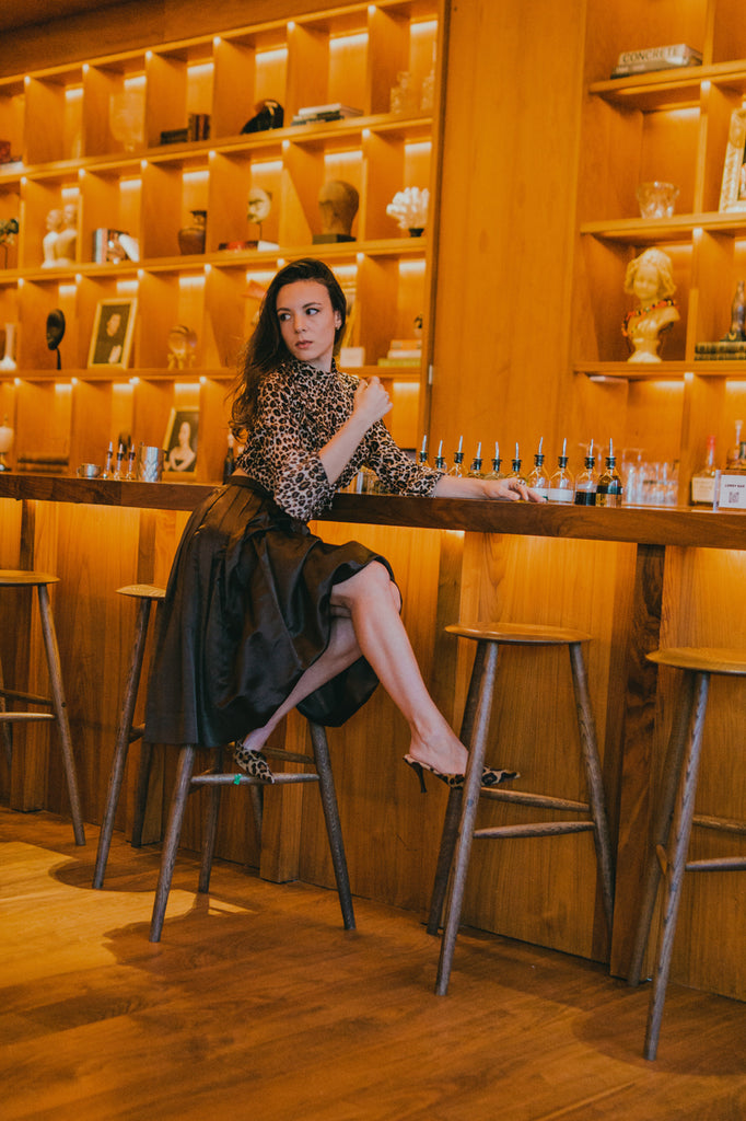 sarah amorelli in leopard print top, brown skirt and leopard print heels sitting at a bar.