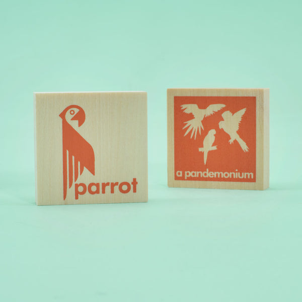 two wooden chips, a parrot is printed on one along with an image of a parrot. The other chip has multiple images of parrots, with the words "a pandamonium." Both images are orange illustrations.