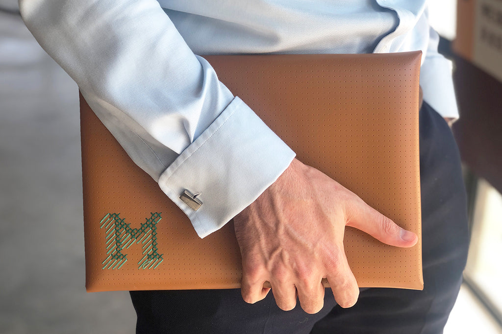 man carrying brown stitched laptop case with M 3D initials stitched in green and teal