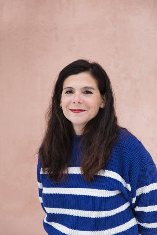 Portrait of the author, Cinthia Vallet - a white woman with long brunette hair, wearing a blue and white striped sweater against a neutral backdrop