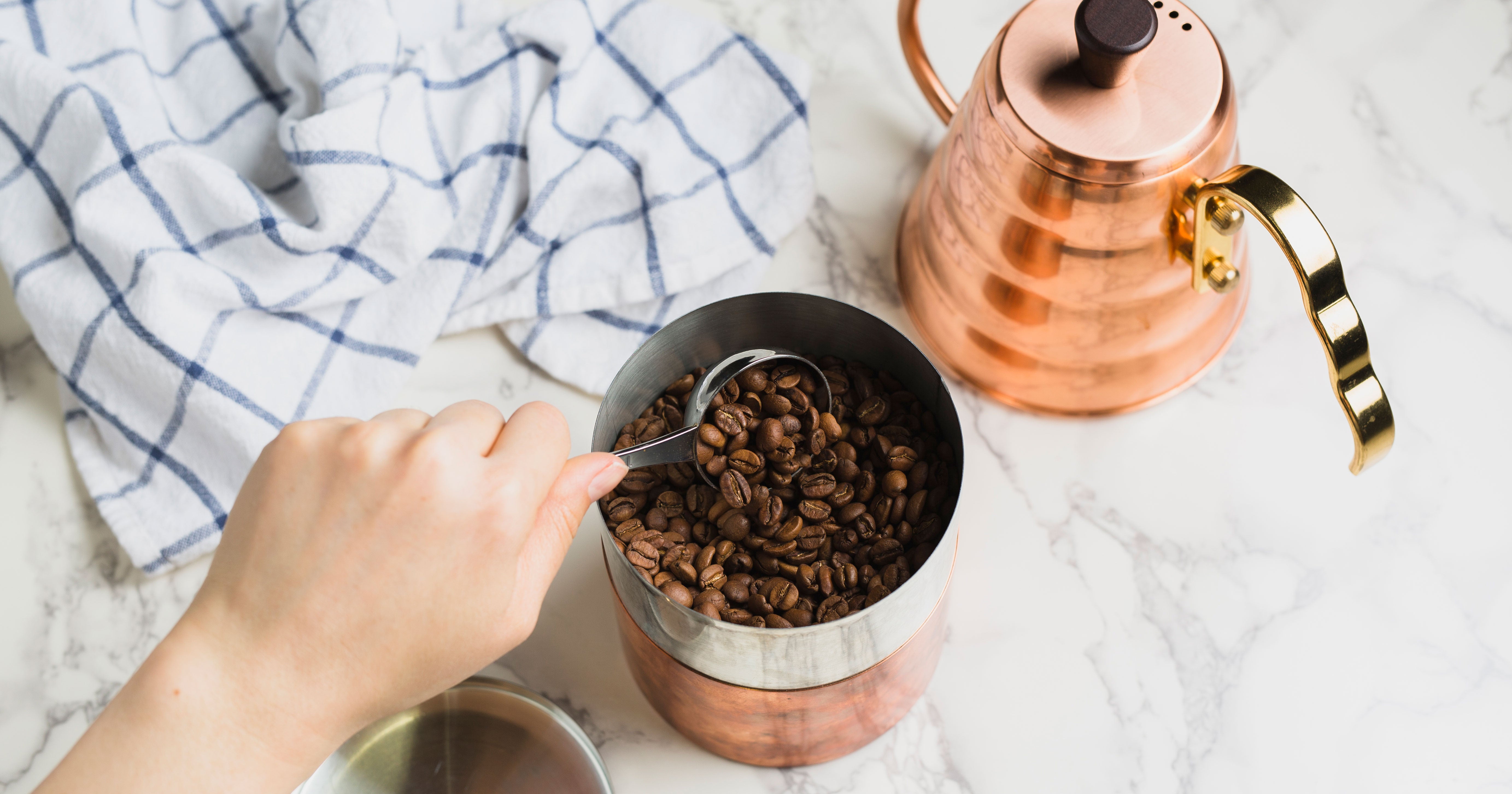 a hand scooping coffee out of a copper canister while a copper hario v60 kettle sits behind it