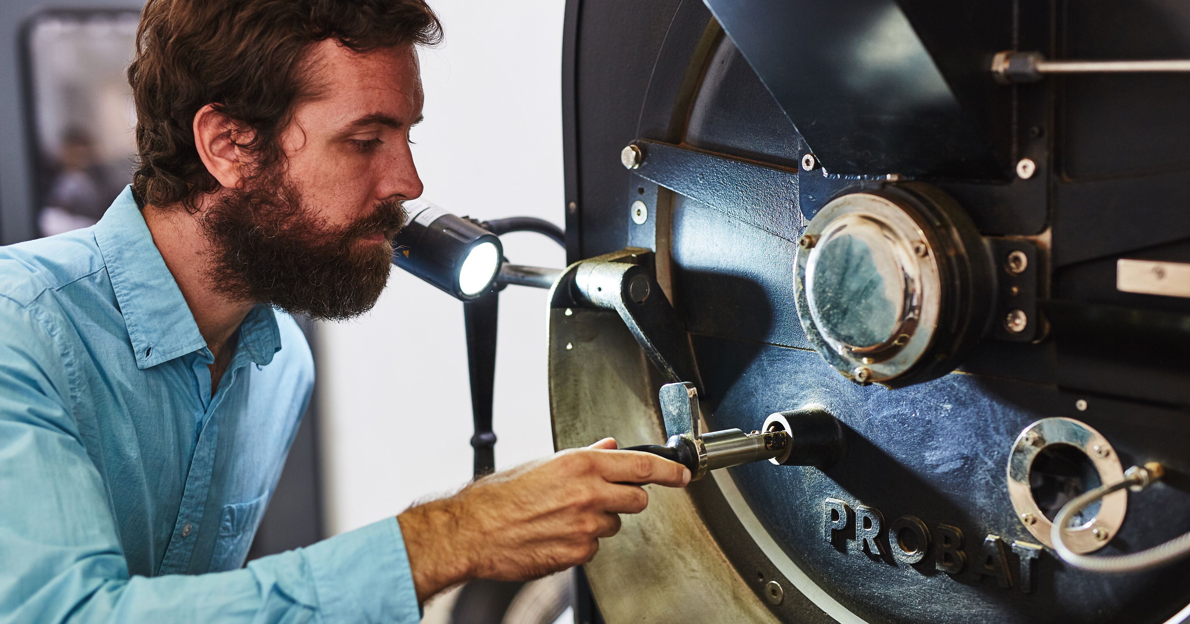 a bearded man in front of a probat roaster inspecting coffee in the trier