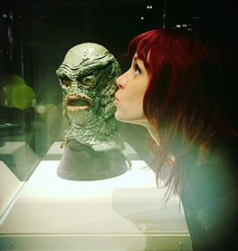 Picture of a red head woman artist kissing a sculpture of the creature from the black lagoon