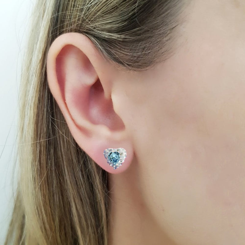 Dainty heart stud earring with an aquamarine heart crystal in the centre (the birthstone for March), on a women ear. The heart shaped stud earring in sterling silver for pierced ears, with a light blue central heart crystal and tiny moonlight crystals around it, in a pave style heart earring, made in Ireland by Lavinia of Magpie Gems Jewellery in Cork.