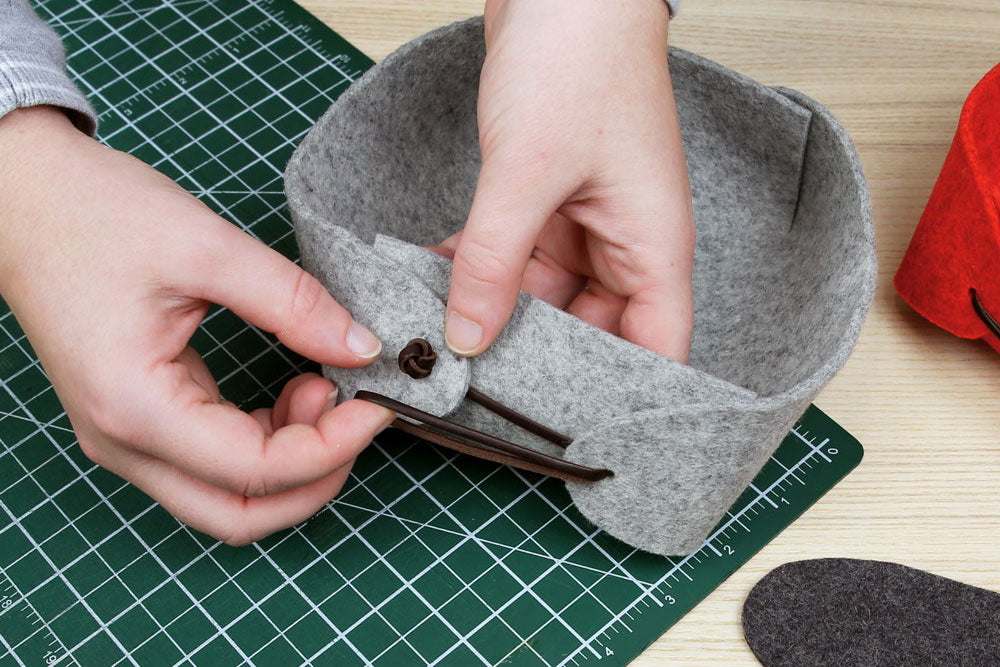A pair of hands making a felt box. They hold the side of the box and a loop of leather cord that they are about to loop around a leather knot.