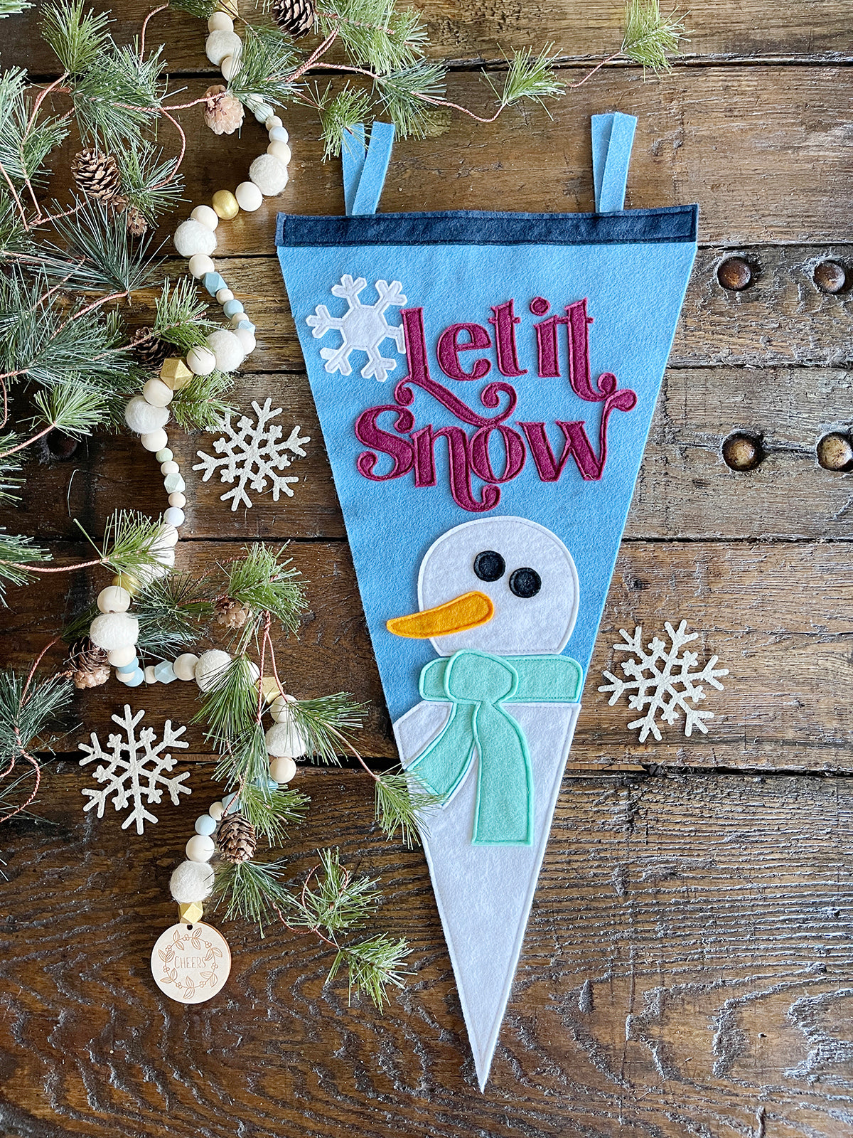 Bird's eye view of a Light Blue felt pennant with red lettering spelling 'Let it snow' and a white snowman with a mint scarf at the bottom. A garland of felt balls and wood beads in neutral colors is laid on pine boughs in the left of the photo.