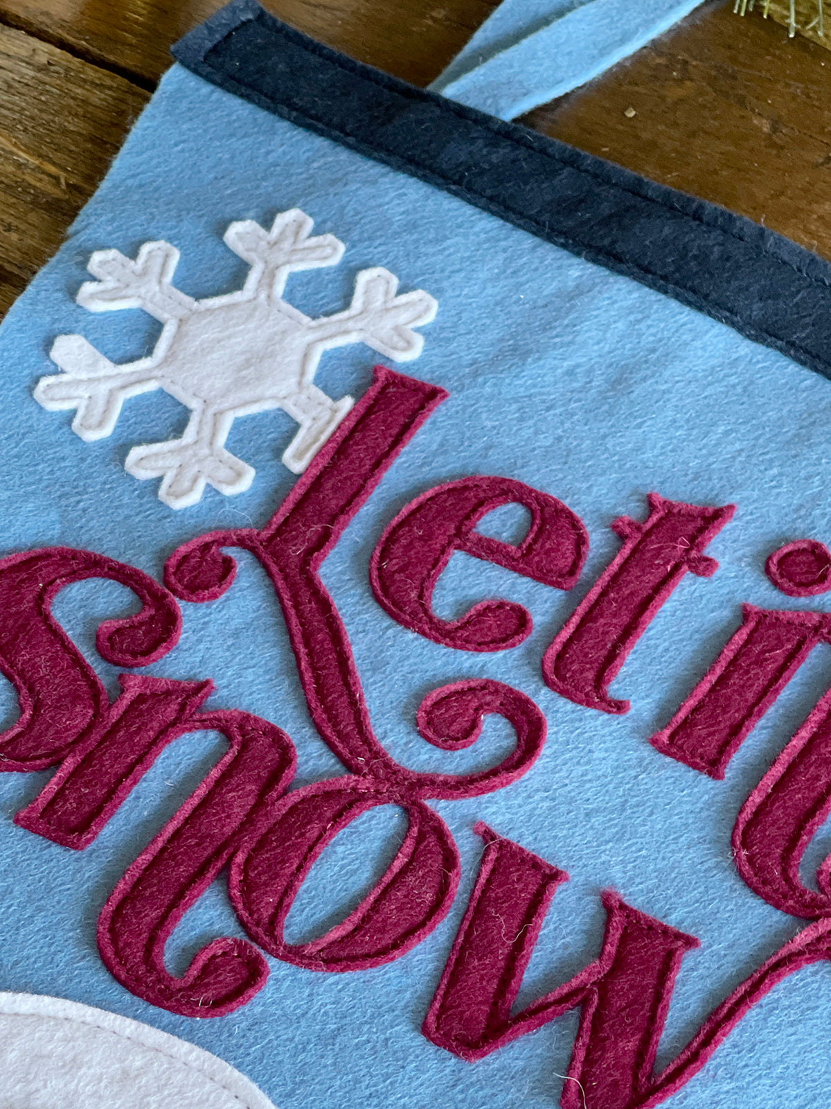A close up photos of red felt lettering spelling 'Let it snow' and a white snowflake sewn onto a light blue background.