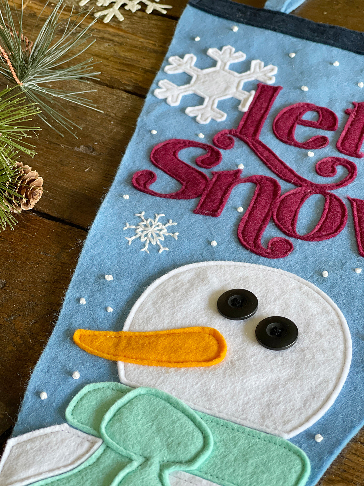 A close up photo of a light blue felt pennant on a wooden table. The pennant has red lettering spelling 'Let it snow' and a white snowman with a mint scarf sewn to the backing. Small white beads have been sewn to the backing.