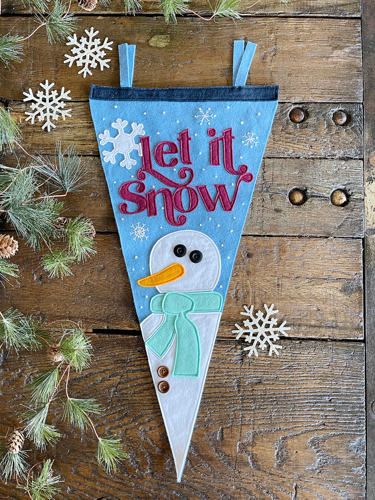 Bird's eye view of a felt pennant on a wooden table. Pine boughs are strewn across the left side of the photo. The pennant is light blue with red lettering spelling 'Let it snow'. A white snowman with a mint scarf sits at the bottom of the pennant. Small white beads have been sewn to the pennant backing.