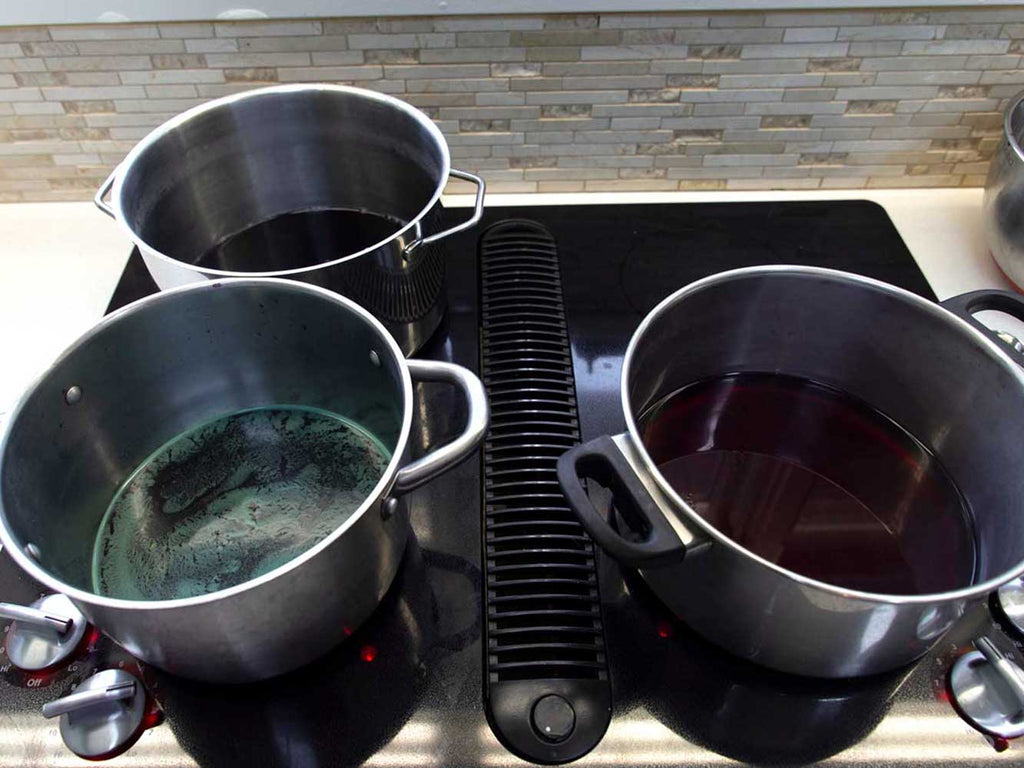 Three stainless steel pots on a black stovetop. Each pot contains red cabbage dye water.