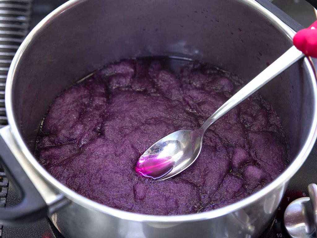 A close up of a pot on a stovetop. Inside the pot is purple carded wool sitting in a red cabbage dye water bath. The wool is being pressed down by a metal spoon.