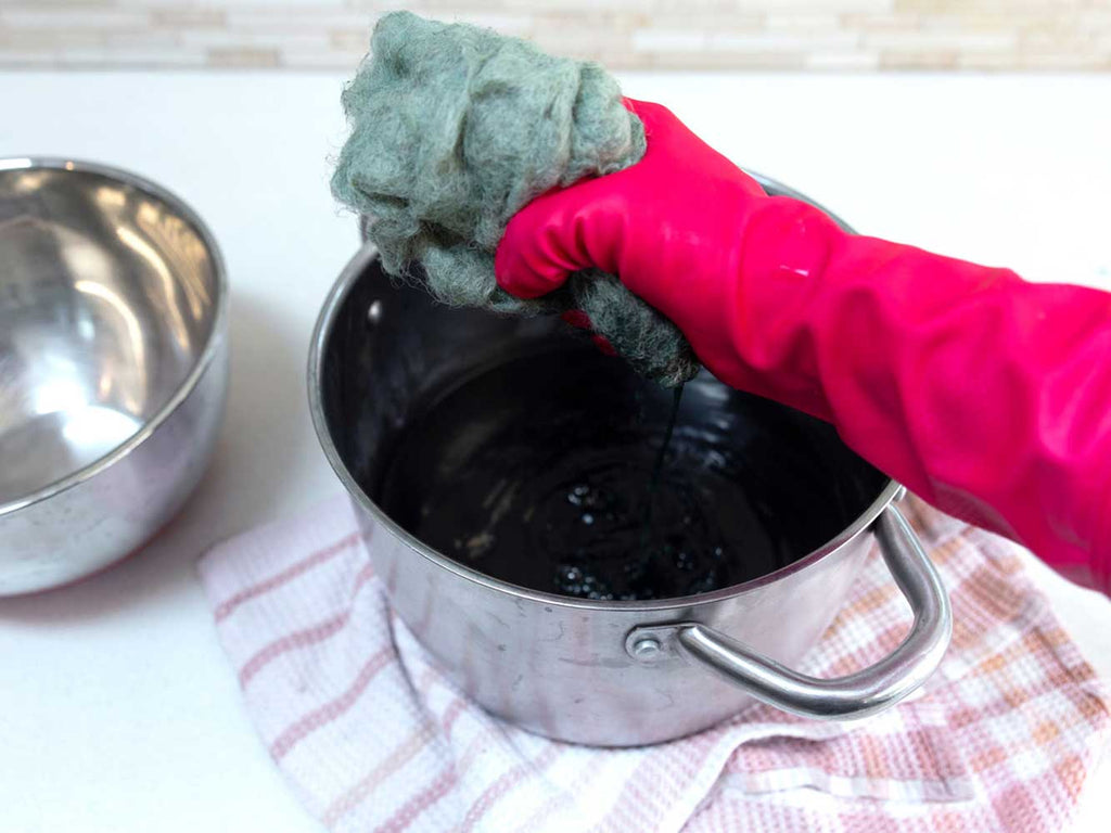 A red gloved hand is squeezing liquid from green carded wool. There is a stainless steel pot on a tea towel underneath the hand catching the liquid.