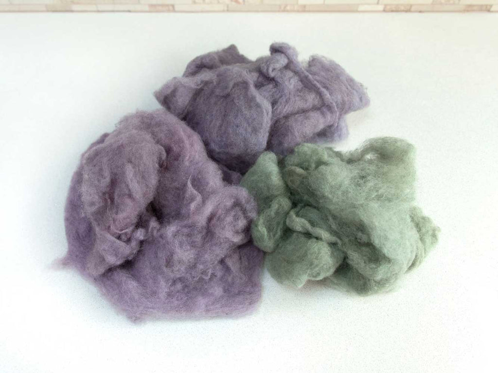 Three piles of fluffy carded wool on a white countertop. One pile is green, one pile is grey-purple, one pile is pink-purple.