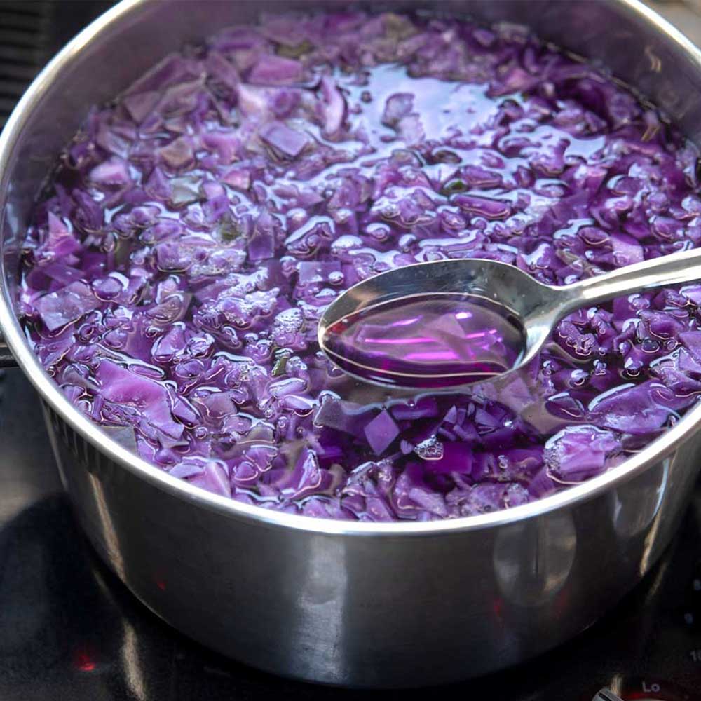 A close up of a stainless steel pot filled with chopped red cabbage and water. There is a spoon filled with the water from the pot in the foreground. The water in the spoon is a pinkish-purple.