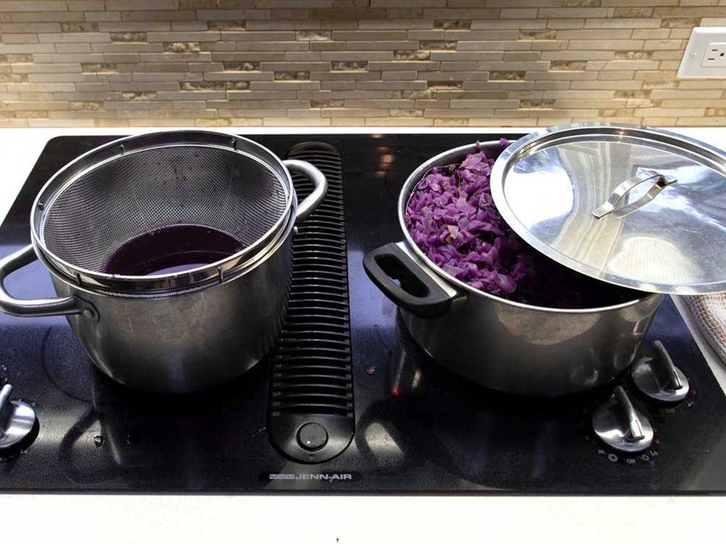 two stainless steel pots sitting on a stovetop. Inside the left pot is a strainer filled with purple water. Inside the right pot is chopped red cabbage that has had the water strained from it.