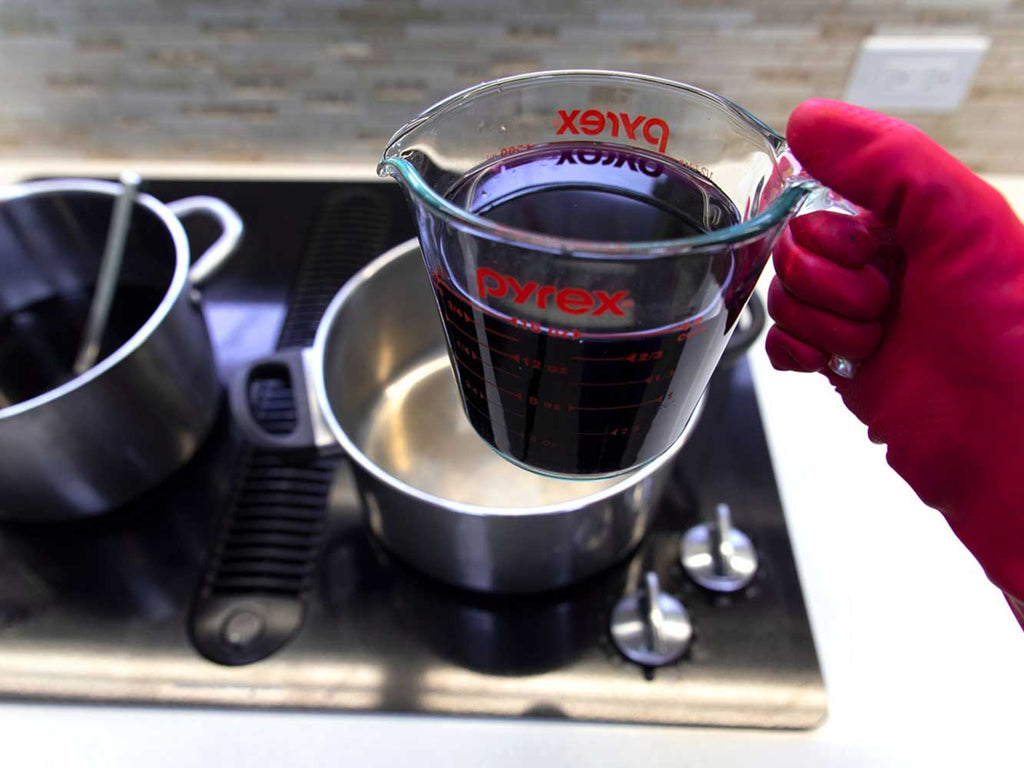 A red-gloved hand holding a measuring cup filled with dark purple dye water. In the background there are two pots sitting on a stovetop.