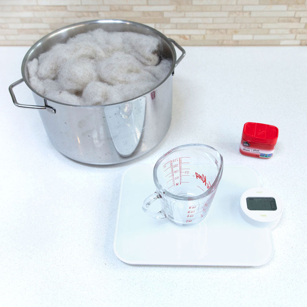 A picture with a measuring cup on a kitchen scale. A spice container of alum and a pot with carded wool is in the background