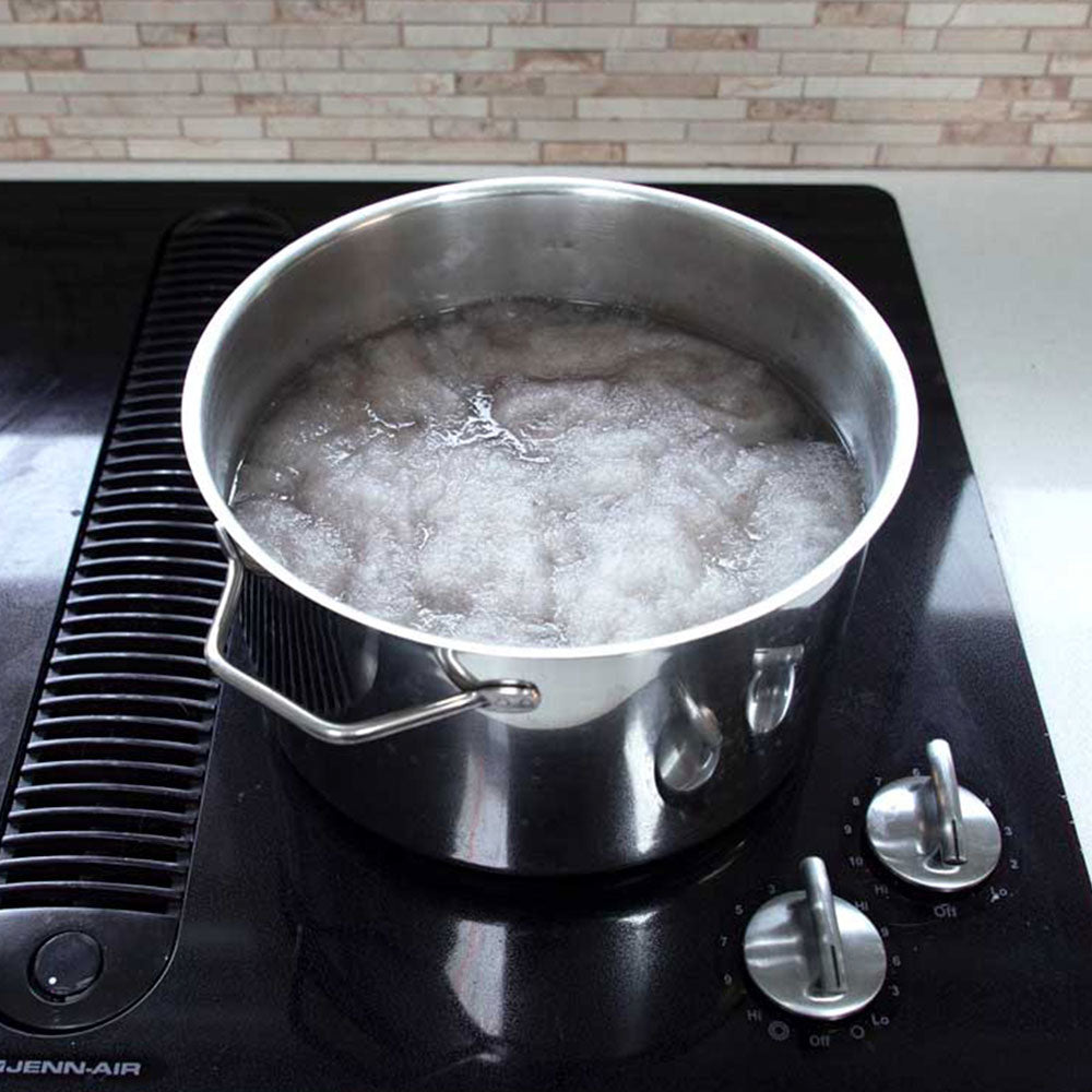 A stainless steel pot filled with carded wool in a warm water Alum solution sitting on a stovetop.