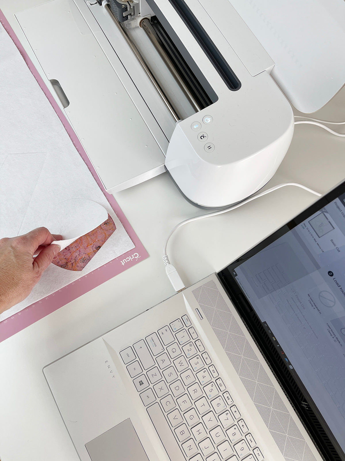 Bird's eye view of a laptop and Cricut machine, a hand is peeling felt away from a Cricut fabric mat