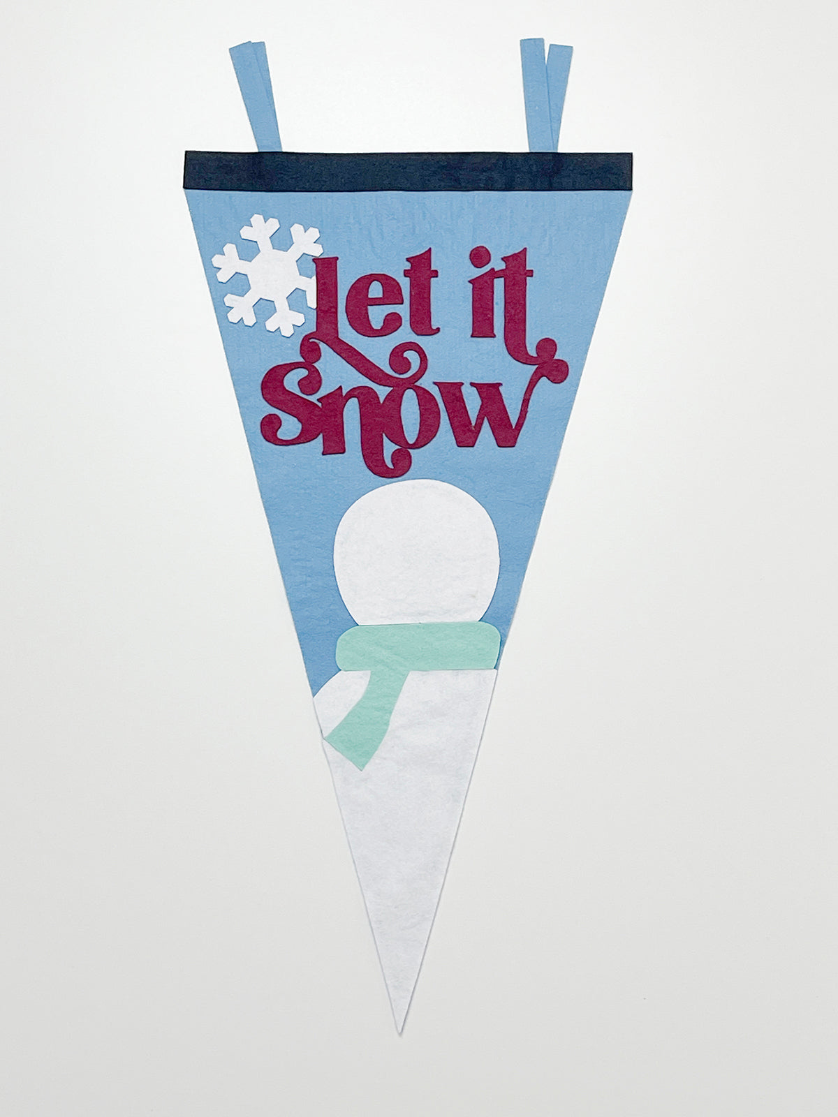 Bird's eye view of felt pennant under construction. Red lettering spells out 'Let it snow'. A white snowman with a mint green scarf is at the bottom of the pennant. The snowman has no facial features.