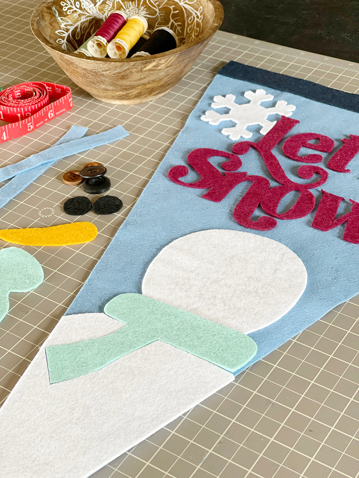 Angled view of felt pennant under construction. Red lettering partially out of frame spells out 'Let snow'. A white snowman with a mint scarf is at the bottom of the pennant. There are felt scraps and buttons to the left of the photo.