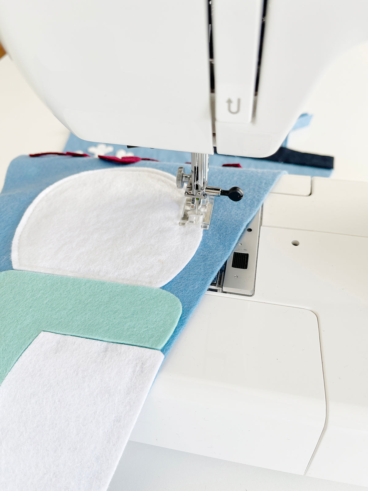 A white felt snowman on a light blue felt background going through a sewing machine.